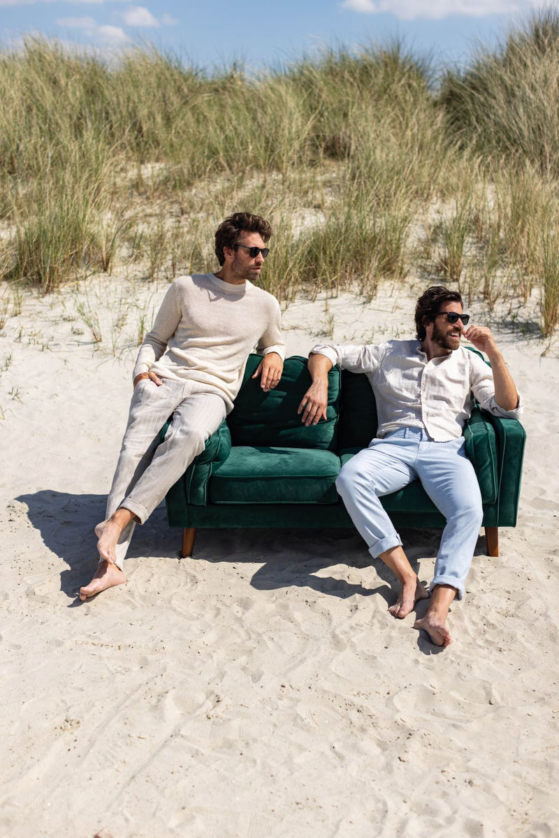 Ein grünes Samtsofa mit modernen, geraden Armlehnen auf einem Sandstrand mit zwei entspannten Männern, die Sonnenbrillen tragen und in einem hellen, lockeren Stil gekleidet sind, umgeben von Dünen und Himmel, symbolisiert Komfort und Ruhe.