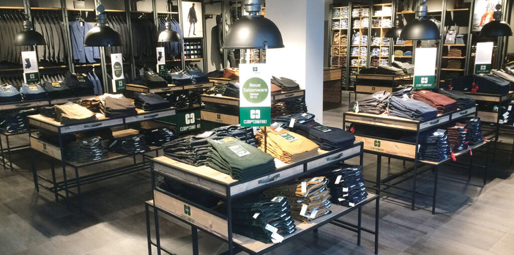 Blick in das Innere eines Modegeschäfts, in dem mehrere schwarzergundene Tische mit gestapelten Kindermodeartikeln wie Hemden, Hosen und Jacken stehen. Im Hintergrund sind Regale mit weiteren Kleidungsstücken sichtbar, die in einem gut beleuchteten Raum sortiert sind.