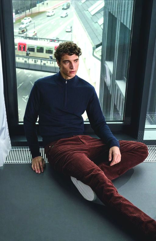 Junge Mann sitzt an einem großen Fenster im Indoor-Bereich, schaut nach draußen, trägt ein dunkelblaues Langarmshirt, braune Cordhose und weiße Sneakers. Hinter ihm ist eine Stadtstraße mit Autos, einer Straßenbahn und Gebäuden sichtbar.