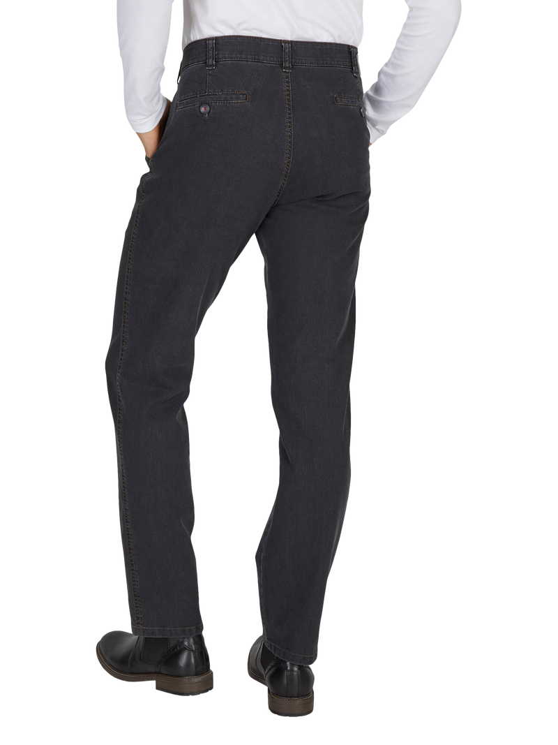 Blick auf die Rückseite einer Person, die dunkelgraue Jeans trägt. Die Jeans hat einen hohen Bund, Seitentaschen und wird mit einem weißen Hemd kombiniert. Die Person trägt schwarze Schuhe und steht mit leicht gespreizten Beinen, die Hände sind in den Jackentaschen.