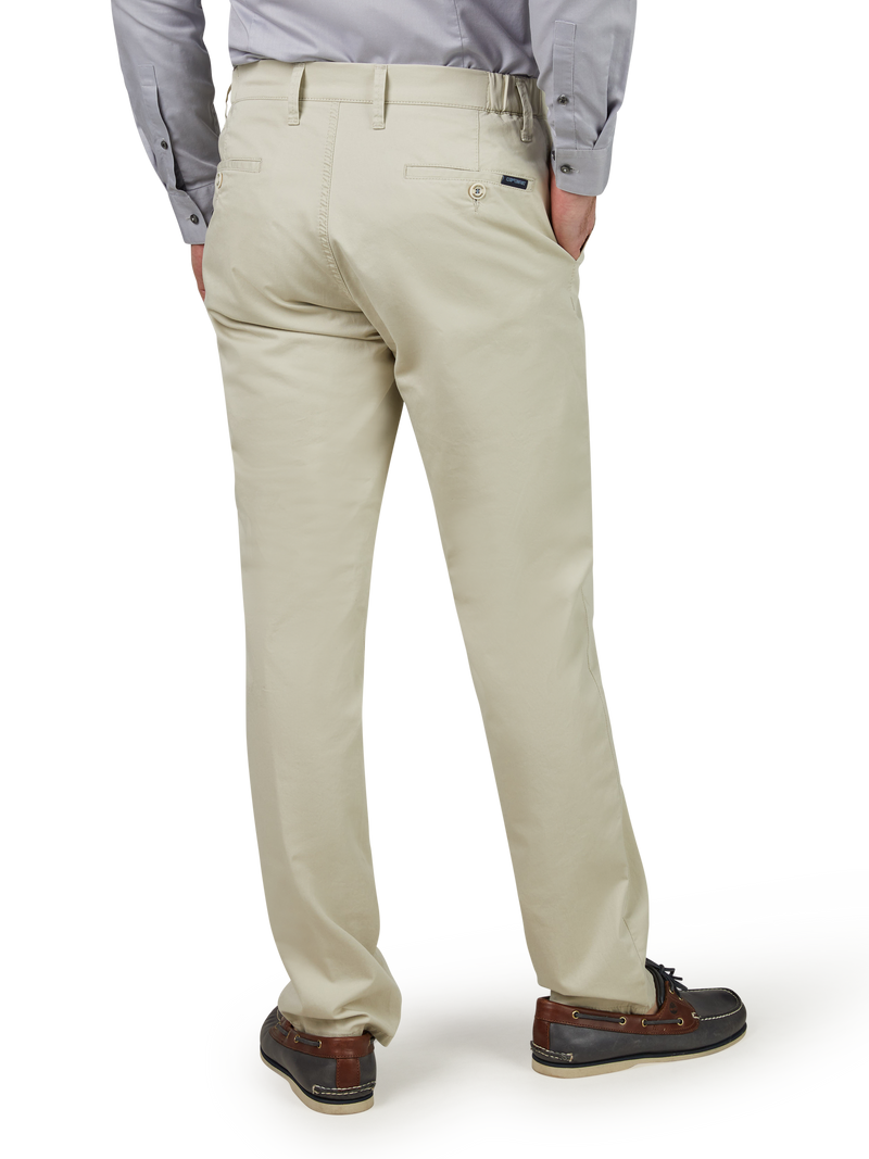 Ein Mann trägt beige Chinos, die bis zu den Füßen reichen, mit einem grauen Hemd. Er steht aufrecht, hat eine Hand in der Tasche und trägt braune Lederschuhe.