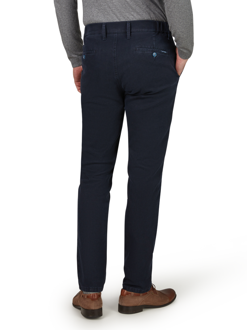 Blick auf die Rückseite eines Mannes, der dunkle, gerade geschnittene Stoffhose trägt. Er hat eine graue Jacke oder Pullover an und trägt braune Schuhe. Seine Hände sind in den Hosentaschen.