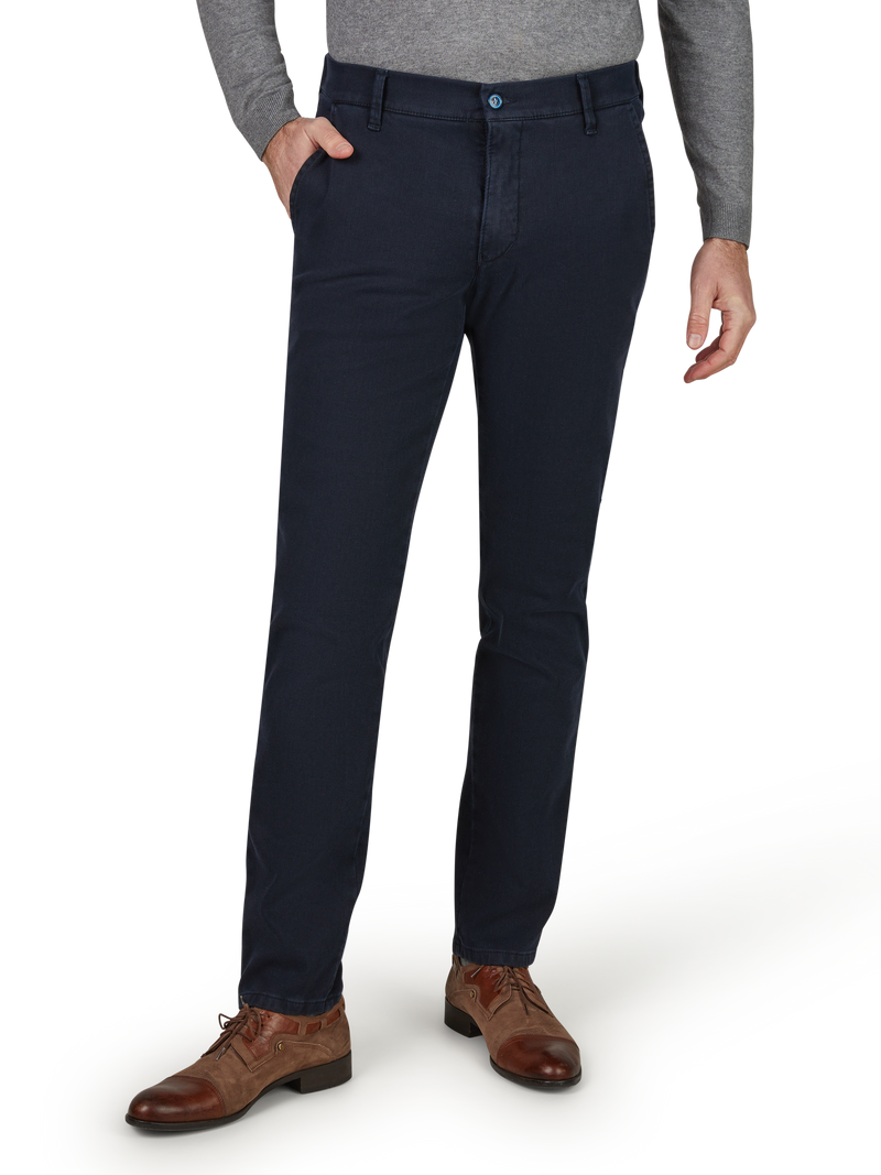 Nahaufnahme eines Mannes in dunklen, gerade geschnittenen Chino-Hosen, grauem Pullover, braunen Lederschuhen, mit einer Hand in der Hosentasche, der vor einem weißen Hintergrund steht.