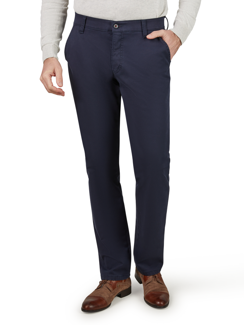 Hosenmodell in dunkelblauer Farbe, gerade geschnitten, mit sichtbarem Reißverschluss und Knopf vorne. Die Person hat eine Hand in der Hosentasche, trägt braune Lederschuhe und ein hellgraues Oberteil.