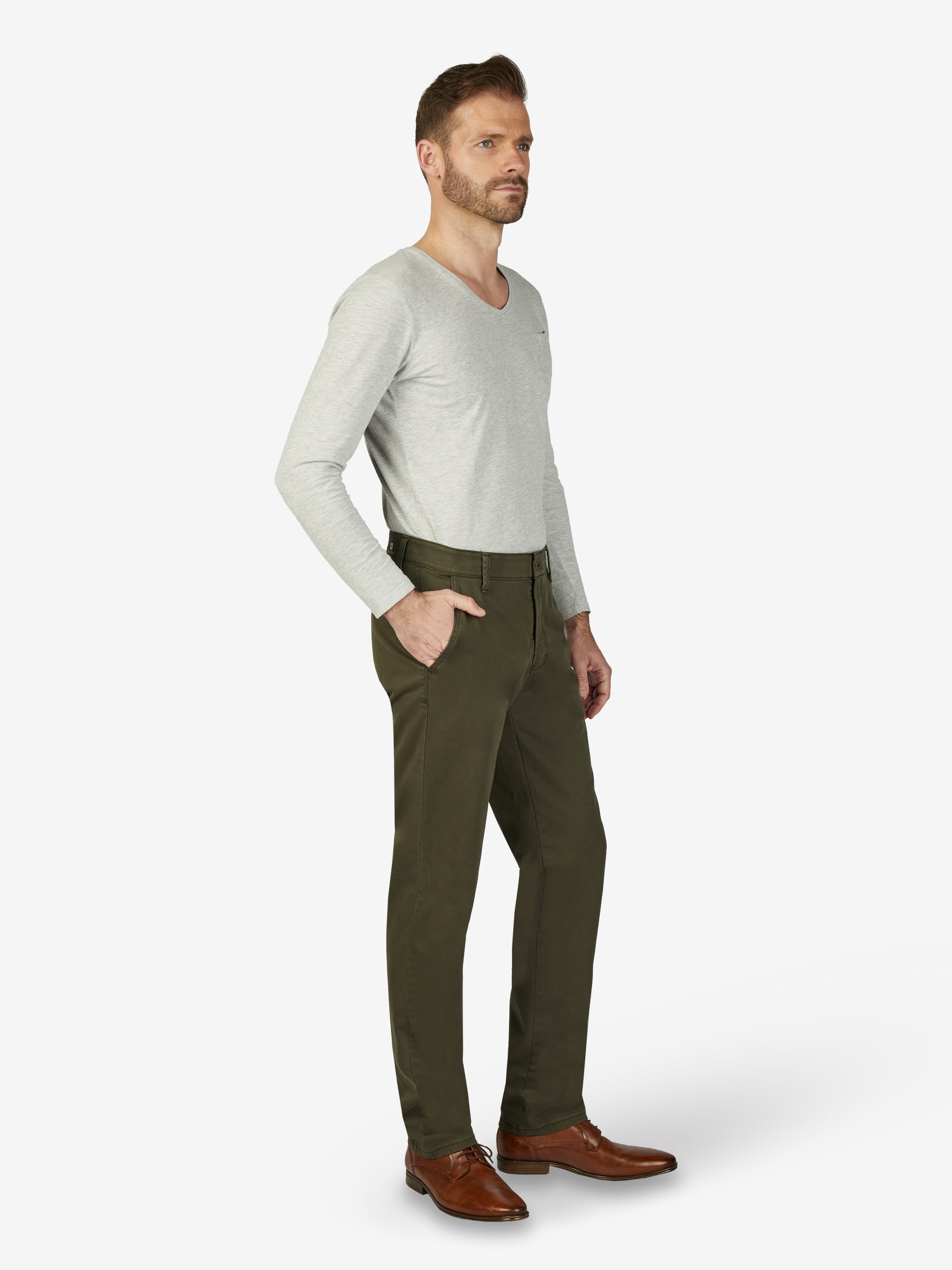 Mann trägt dunkel khakifarbene GARVEY Flat Front-Hose aus elastischem Baumwoll-Gabardine mit High Stretch, Komfortbund, Hemdenstopper und schmaler Fußweite, kombiniert mit hellem Pullover und braunen Lederschuhen, ideal für Herbst/Winter.