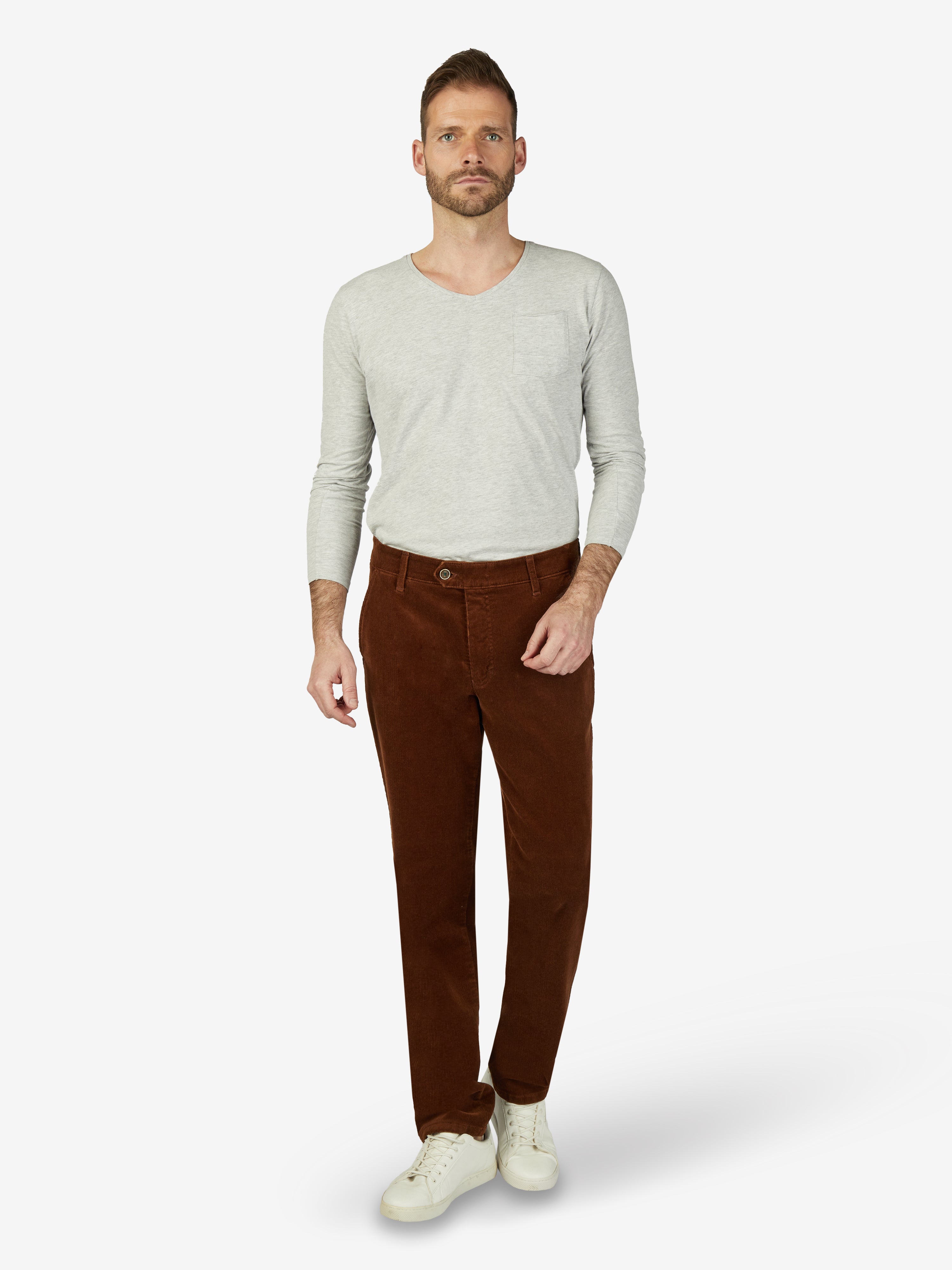 Mann trägt eine braune GARVEY 8200 Flat Front-Hose aus Feincord mit Stretch und Komfortbund, kombiniert mit einem hellgrauen Langarmshirt und weißen Sneakern. Die Hose hat eine normale Leibhöhe, schmalen Beinverlauf, Hemdenstopper, Bundverlängerung mit Knopf und Haken, Made in Green by Oeko-Tex, perfekt für Herbst/Winter.