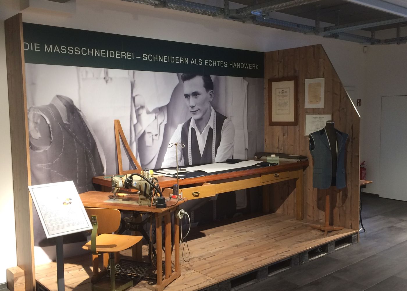 Blick auf eine Ausstellung zum Thema Schneiderei: Im Hintergrund ein großes Schwarz-Weiß-Foto eines Mannes in Schneidereiform, vorne ein langer Holzarbeitstisch mit Nähmaschine und Schnittmustern, rechts ein Kleidungsstück auf einem Mantelständer, links ein gelber Stuhl und ein Info-Schild.