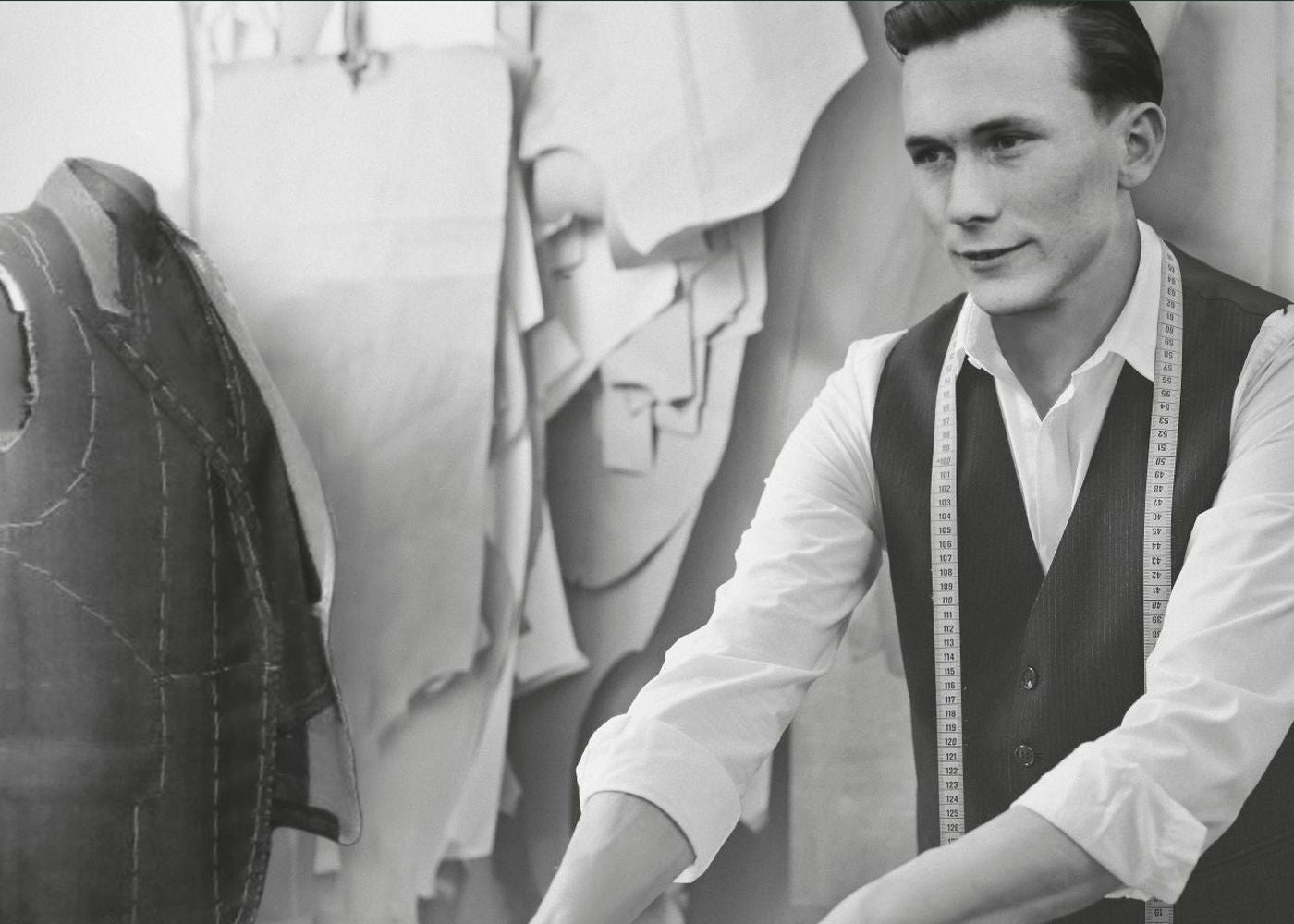 Ein junger Mann mit dunklem, gepflegtem Haar, trägt ein weißes Hemd und eine dunkle Weste. Er sitzt an einem Tisch in einer Schneiderei oder Modewerkstatt, mit einem Maßband um den Hals. Im Hintergrund sind Schnittmuster und Stoffe an der Wand sichtbar.