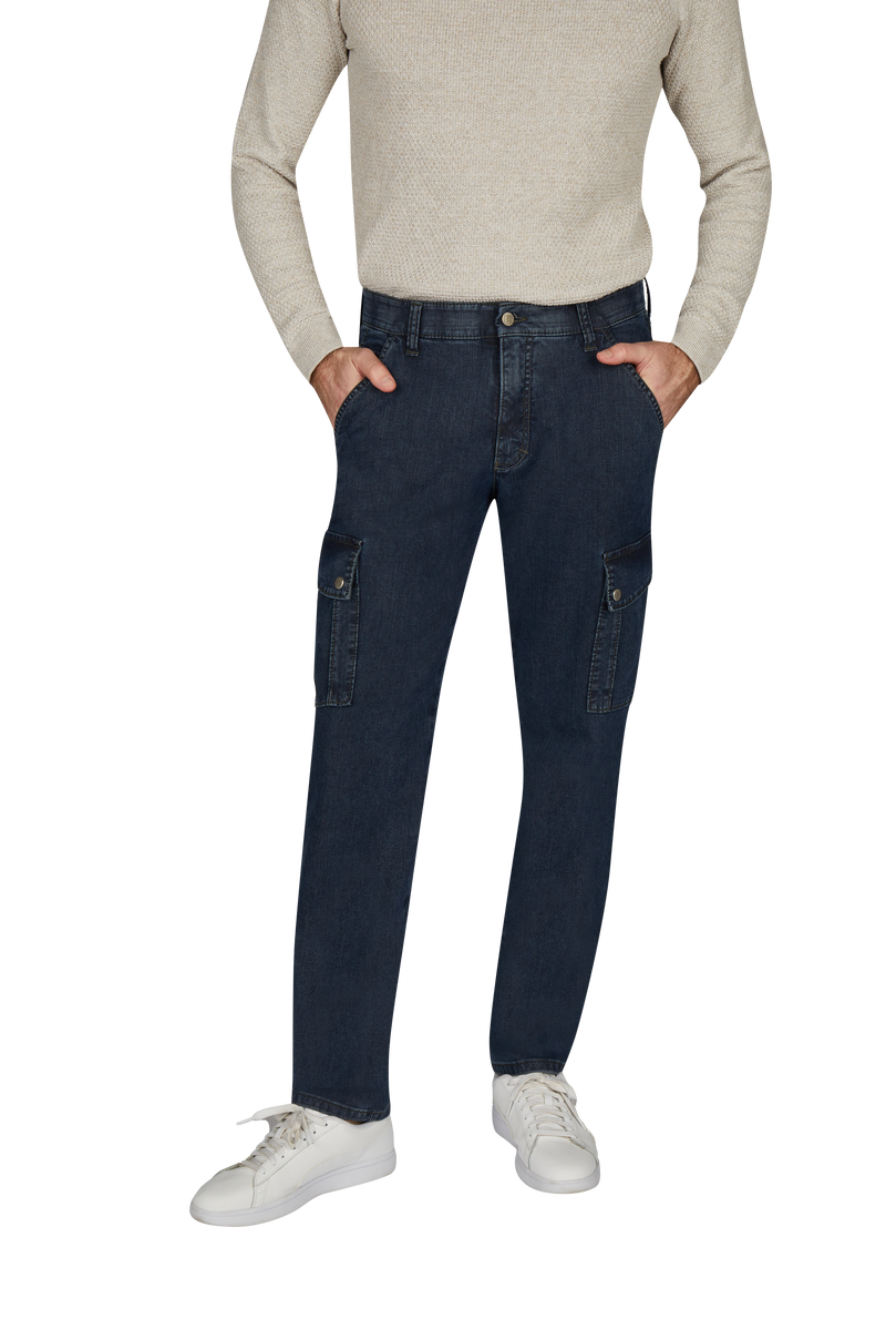 Ein Mann trägt eine beige Pullover, dunkle Cargo-Jeans mit mehreren Taschen und weißen Sneakers, steht mit den Händen in den Taschen.