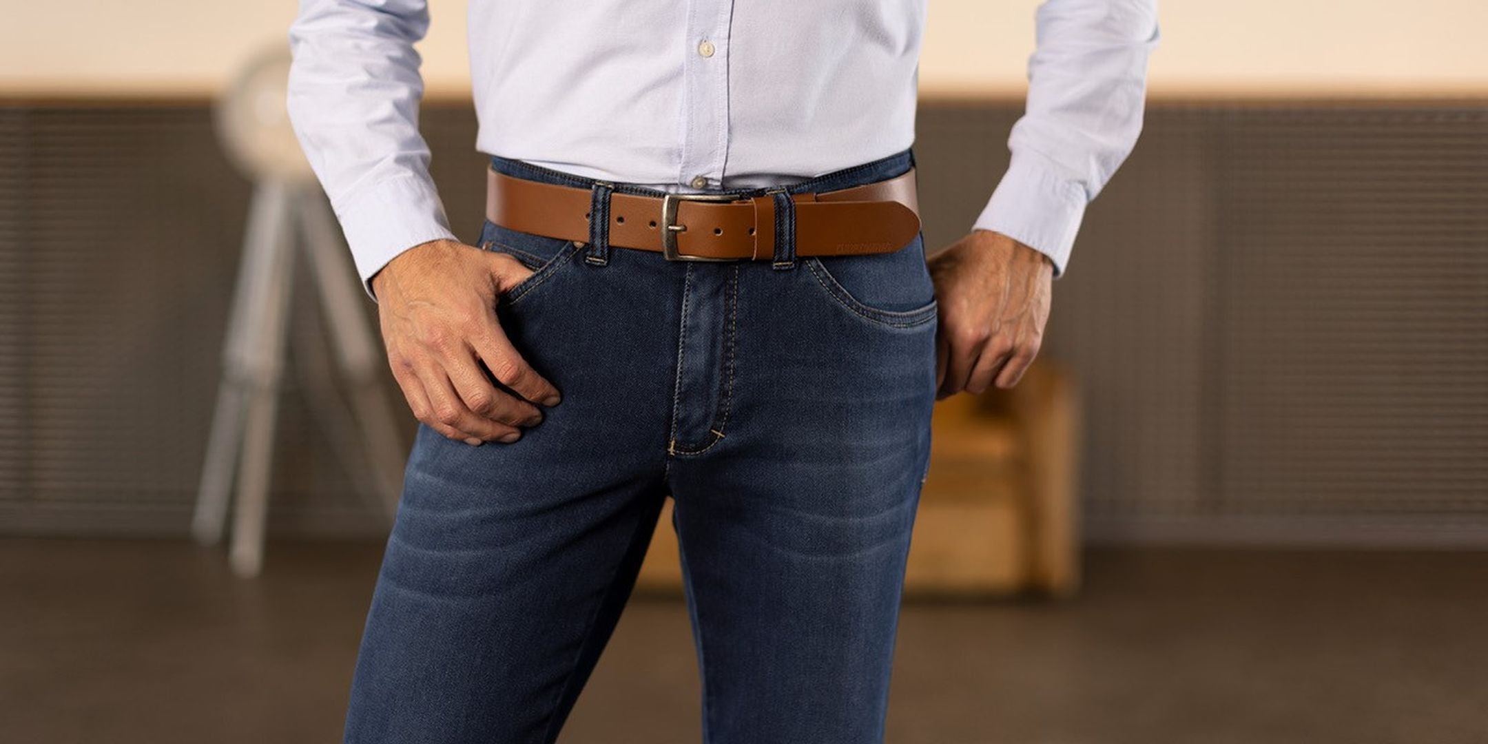Der Bildausschnitt zeigt eine Person, die dunkelblaue Jeans trägt und ihre rechte Hand in die Hosentasche steckt. Die Person trägt ein weißes Hemd und einen braunen Gürtel. Im Hintergrund sind unscharf Möbel und ein Arbeitsplatz sichtbar.