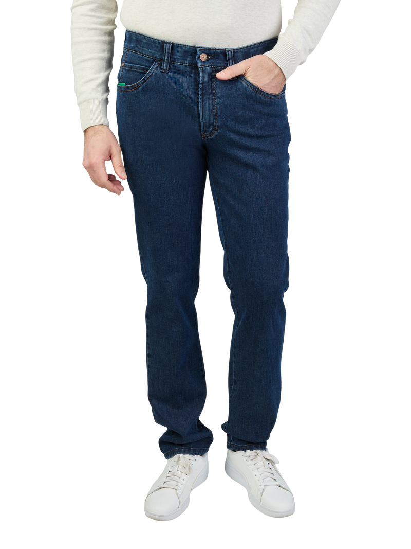 Ein Mann trägt dunkle Jeans, ein weißes Oberteil und weiße Sneakers. Seine rechte Hand liegt in seiner Hosentasche, während seine linke Hand locker an der Seite hängt.